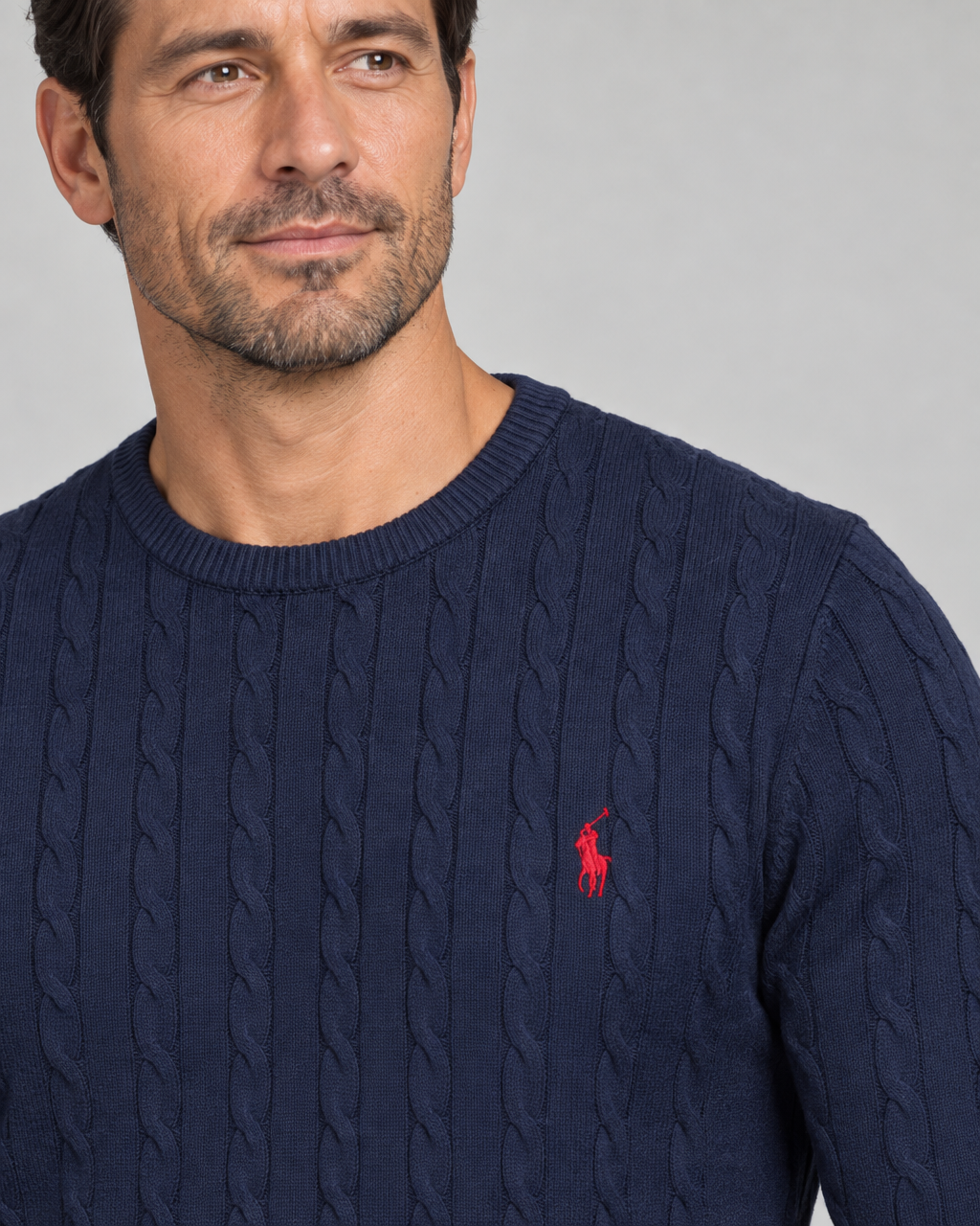 Close-up of a man wearing a dark blue cable-knit crew-neck jumper with red chest embroidery