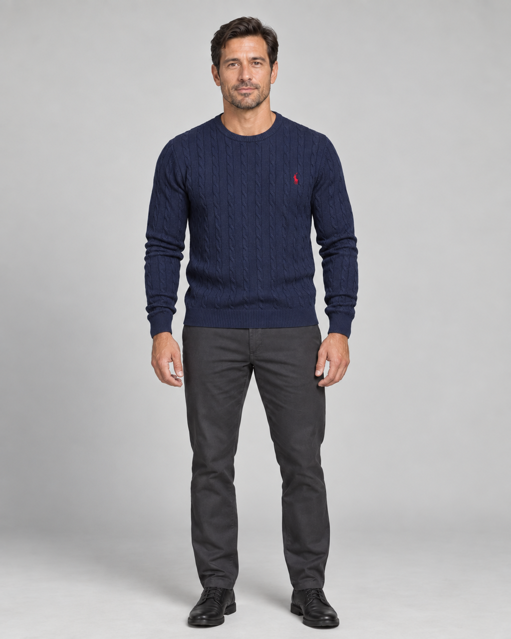 Front full-body view of a man wearing a dark blue cable-knit crew-neck jumper with a small red chest motif.