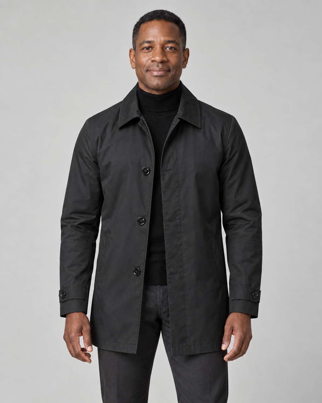 Front cropped view of a man wearing a black thigh-length coat with a pointed collar, buttons, side pockets and cuff tabs.